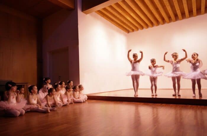 Young dancers on stage demonstrate how dance classes can benefit your child, with poised ballet movements in white tutus, while their attentive peers sit on the floor, embodying the focus and camaraderie fostered through performing arts.