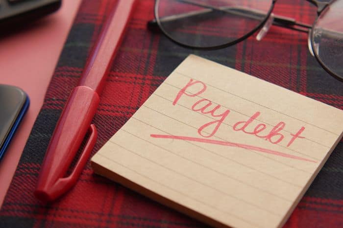 A note pad with the message Pay Debt written on in pink pen surrounded by a pair of glasses and the pen.