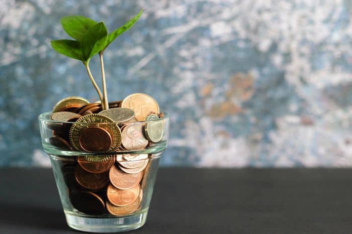 A glass jar saving pot filled with coins and a green sapling growing out of it to symbolise growing savings and manging money better.