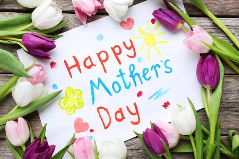A colourful "Happy Mother’s Day" hand-painted sign surrounded by pink, white, and purple tulips on a rustic wooden background, perfect gift inspiration for Mother’s Day.