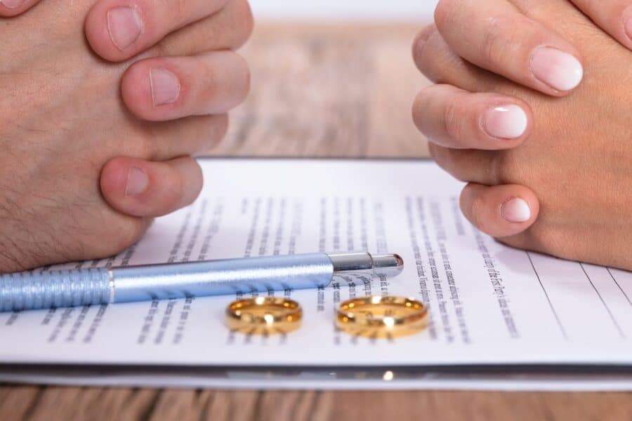 Two pairs of clasped hands positioned opposite each other over a divorce document, with a blue pen and two gold wedding rings placed on the paper.