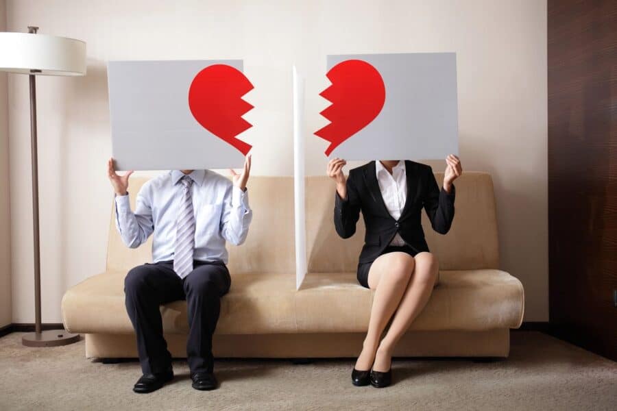 Two individuals sitting on a sofa, separated by a partition, each holding a sign with one half of a broken red heart, symbolising the struggles of dealing with a nasty divorce.