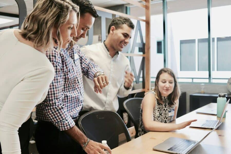A group of four colleagues engaged in a discussion around a laptop in a modern office setting. One person points at the screen while others look on attentively, developing a collaborative work environment to stop employees calling in sick.