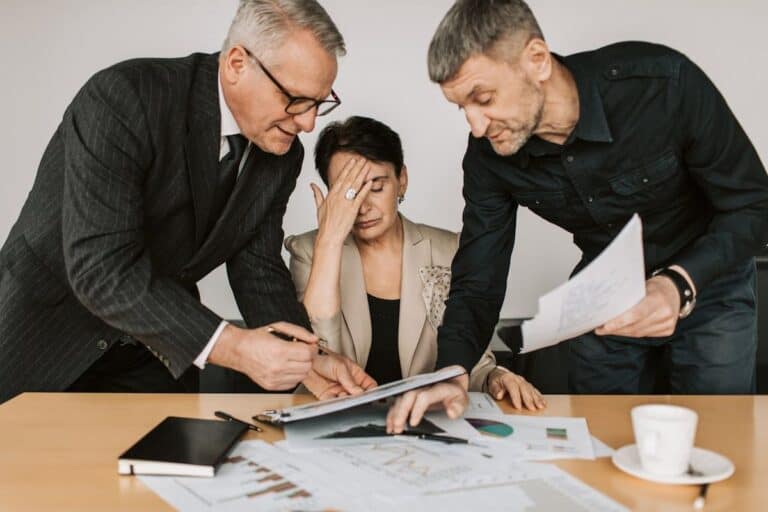 Three colleagues working at a desk filled with documents and charts, looking stressed and overwhelmed. One person has their head in their hand, while the other two lean over to discuss the paperwork. This image highlights the pressure and potential stress in the workplace and needs to be address to stop employees calling in sick.