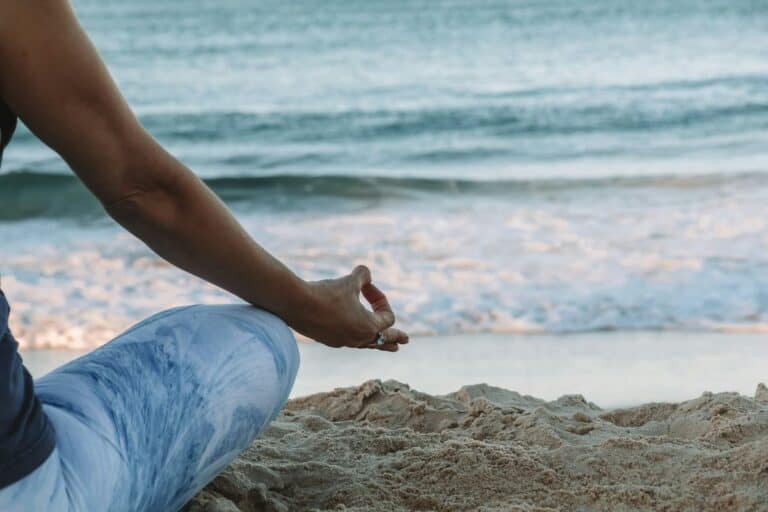 Close-up of a person's side profile in a meditation pose, fingers touching in a mudra, dressed in light blue attire, with a serene beach backdrop highlighting the benefits of living in the present moment.