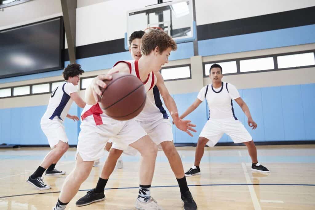 Teenage boys playing basketball in a school gym during a PE lesson, with one student dribbling while others defend.
