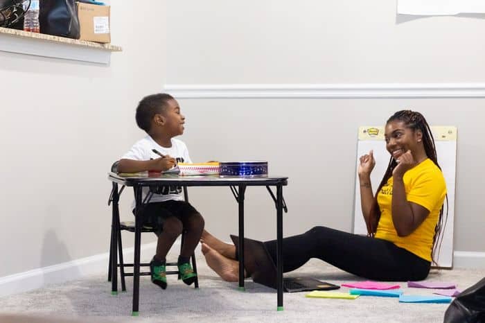 Parent sat on floor praising a child who is sat at a workstation.