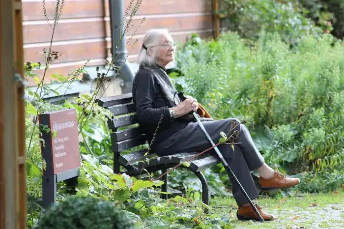 Old woman with a walking stick sat on a bench outside at a nursing home.