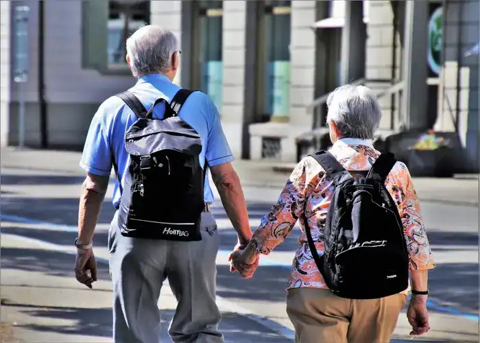 An old couple walking hand in hand wearing travel backpacks.
