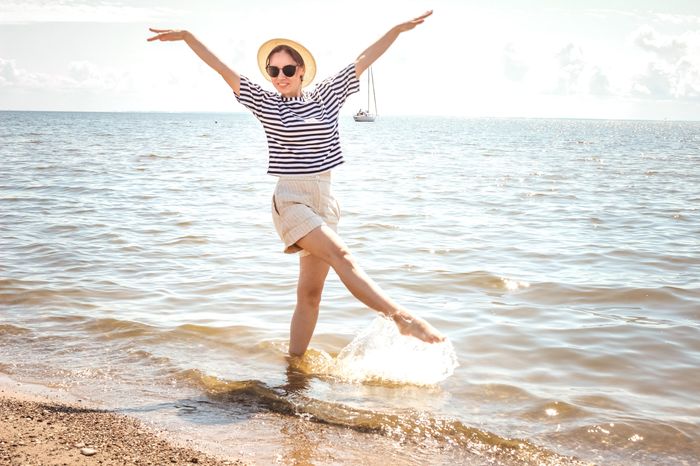 Woman in a striped shirt, beige shorts, and a sun hat joyfully splashing water with one leg at a sunny beach, with a sailboat in the distance.