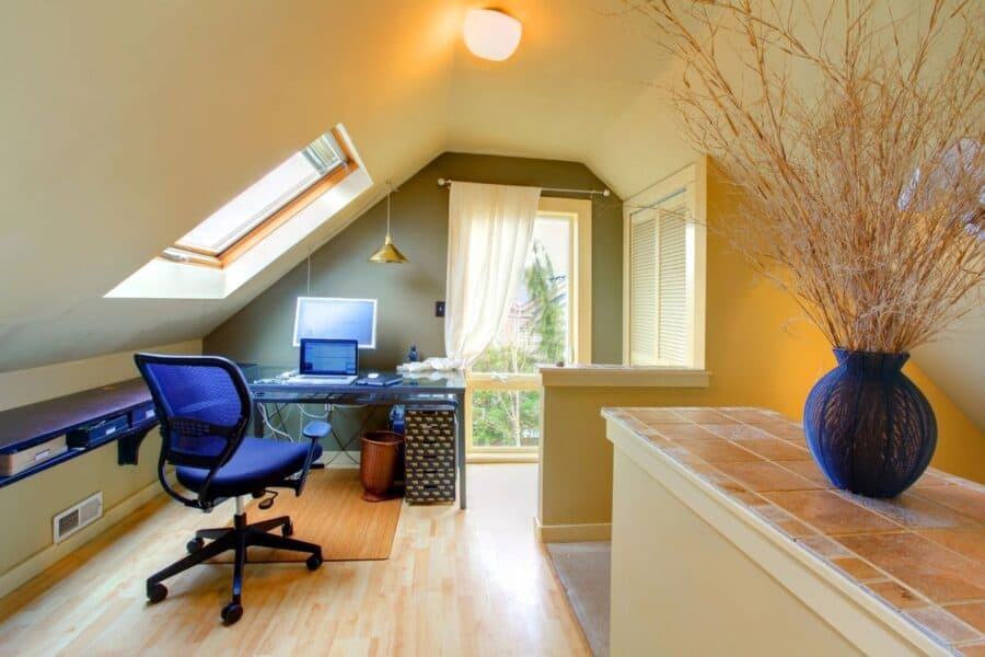 A compact home office setup in a loft with a blue ergonomic office chair at a glass desk holding a computer monitor and accessories. The room features a slanted ceiling with a skylight, warm lighting, and a window with sheer curtains for natural light.