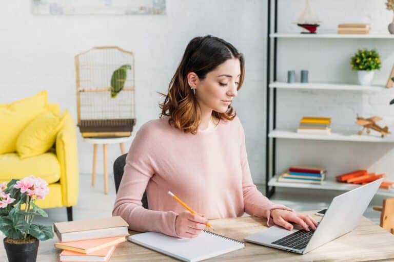A woman in a light pink sweater working at a laptop on a wooden desk with a notepad and pencil in hand. The home office space is bright and organised, featuring a yellow sofa, a parrot in a cage, and a shelf with books and decor.