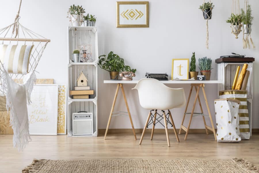 A chic home office featuring a white desk with a modern white chair, surrounded by plants, decorative items, and natural textures like a jute rug and hanging planters. A cosy hammock adds a relaxing touch to the stylish and functional workspace.