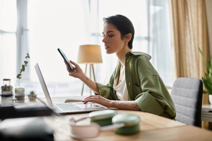 A woman in a green shirt working at a laptop while checking her phone, seated in a well-lit home office with soft natural light streaming through large windows and a warm floor lamp in the background.