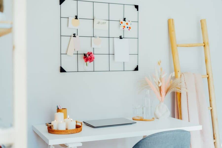 A clean and minimalist desk setup with a white table featuring a laptop, decorative candles in a wicker tray, and pastel stationery. Above the desk is a grid memo board holding notes, cards, and a small vase with flowers, complemented by a wooden ladder and soft pink throw in the background.