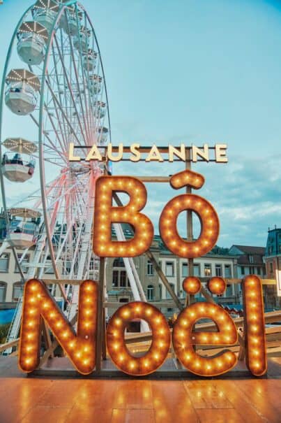Prominent illuminated sign saying 'BÔ Noël' with the word 'Lausanne' above, set against the backdrop of a large Ferris wheel at dusk. The scene captures the festive spirit of the Christmas market in Lausanne, Switzerland, with the sign's large glowing bulbs creating a warm and inviting atmosphere.
