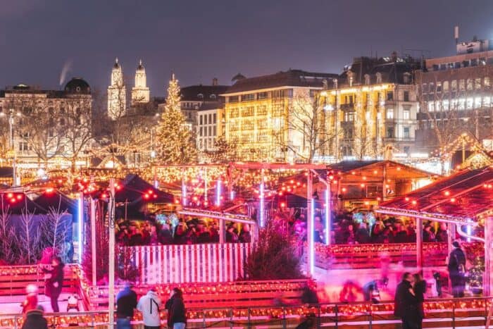 A bustling Christmas market in Zurich, with festive lights illuminating rows of wooden stalls selling holiday crafts and seasonal treats. Crowds of people, bundled in winter attire, stroll through the market, enjoying the festive atmosphere.