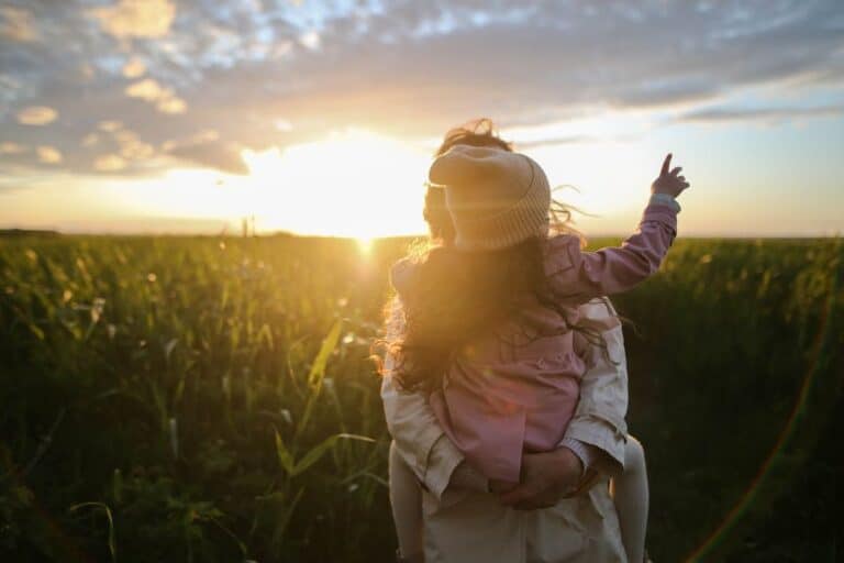 A child in a pink jacket and cream beanie is lifted against a sunset backdrop over a lush field, symbolizing new beginnings with the inspirational theme Change Your Life and be Happier.