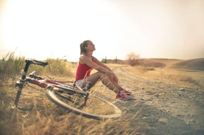 A woman in a red tank top and shorts takes a break beside her lying bicycle, enjoying a moment of rest on a dusty country road with the soft glow of sunset in the background.