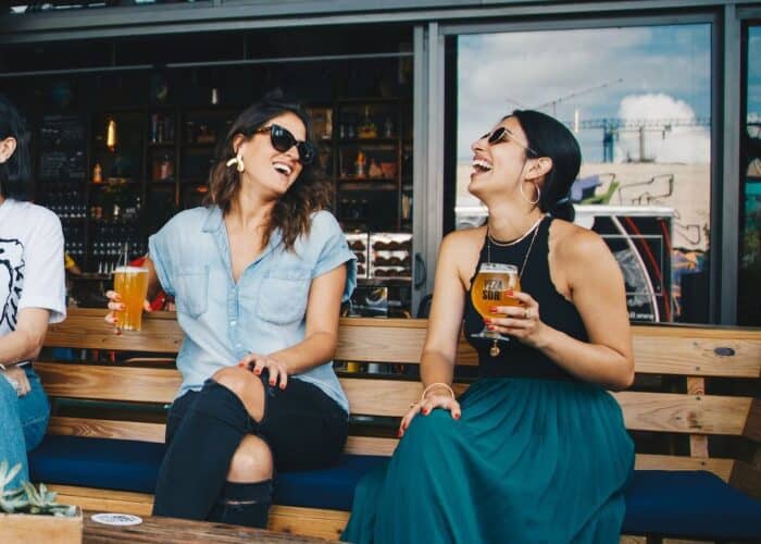 Two women sharing a joyous moment at an outdoor brewery, one holding a glass of beer, both laughing heartily in casual attire, embodying a relaxed and cheerful ambiance.