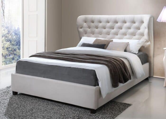 An elegant bedroom setting with a large, tufted headboard on a white upholstered bed, layered with crisp white sheets and a plush brown blanket. The room has a soft grey shag rug, a glimpse of a snowy landscape through the window and a soft glow from a lamp with a lavender shade, creating a serene and luxurious atmosphere.
