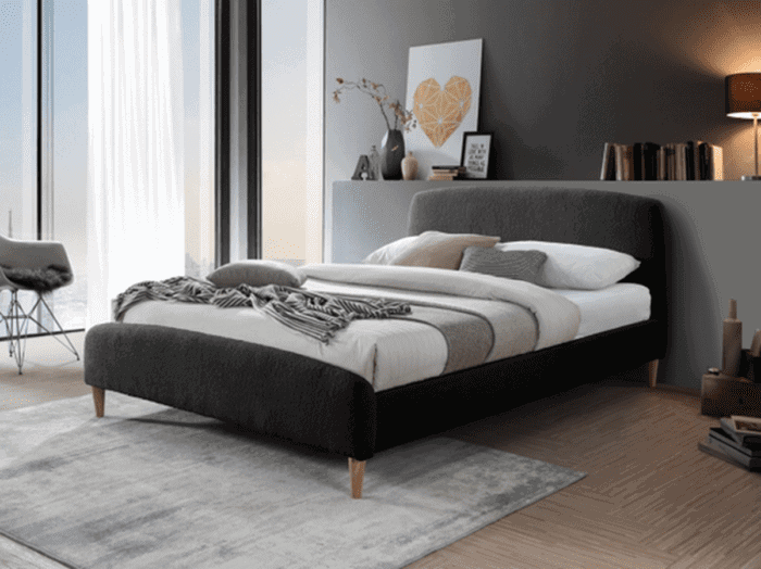 A minimalist bedroom with a dark grey upholstered bed on a light grey area rug, complemented by white and beige bedding and a striped throw. The room is decorated with a geometric yellow artwork, a vase with cherry blossoms and a dark shaded table lamp, all against a grey wall, with natural light streaming in through sheer curtains.
