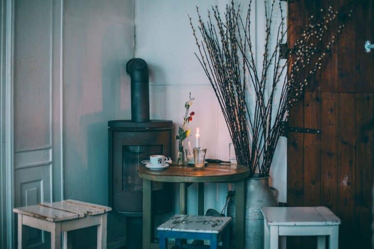 A cosy rustic setting with a French country interior design influence, featuring a wood-burning stove against a muted wall, a wooden table with a teacup, a lit candle and fresh flowers, surrounded by distressed stools and a large metal milk jug filled with willow branches, all against the backdrop of a wooden barn door.
