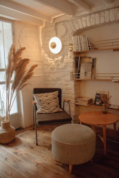 A serene nook with a vintage charm in a room with French country interior design elements, featuring a textured white brick wall with a round wall lamp, a cosy armchair with a geometric-patterned pillow, a straw basket with pampas grass, wooden floor shelving with assorted books, a round wooden table and a patterned ottoman on warm wooden flooring.