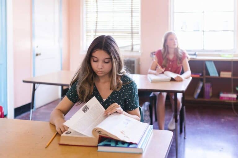 Two teenage students in a bright classroom setting, with the foreground showing a focused girl reading a textbook and the background featuring another student taking notes.