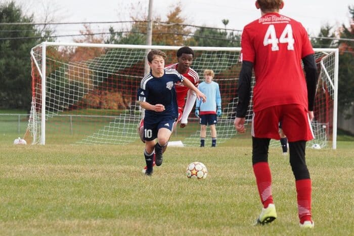 A teenage soccer player in a navy blue jersey, marked with number 10, skillfully dribbles a soccer ball on a grassy field, with opponents in red jerseys and a goalpost in the background. This image represents how engaging in hobbies like soccer can support teenagers in overcoming academic challenges by providing a healthy outlet for stress.