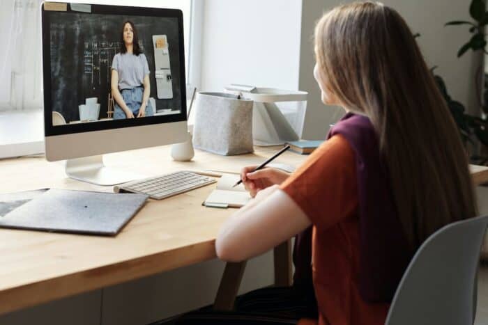 A teenage girl in a rust-coloured top sits at a wooden desk taking notes from an online tutoring session; the tutor, a woman in a casual t-shirt, appears on the large desktop monitor against a blackboard background, during remote education.