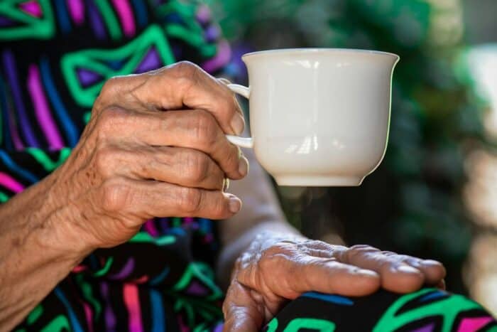 Close-up of an elderly person's hands holding a white ceramic cup, with a colourful patterned garment in the background, symbolising warmth and care in a senior living environment.