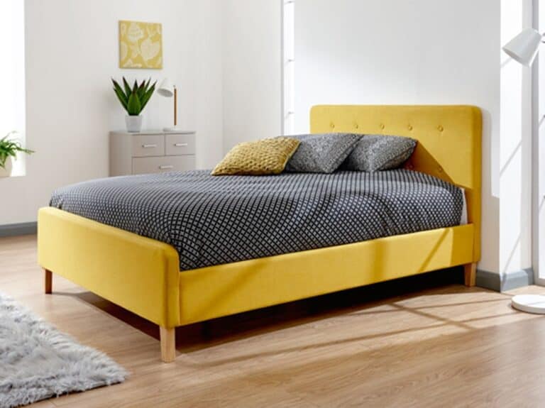 A modern bedroom following the new neutrals in interior design theme, featuring a mustard yellow upholstered bed with a buttoned headboard, dressed in geometric-patterned bedding. A grey bedside drawer, decorative plant and framed artwork complement the clean, bright interior.