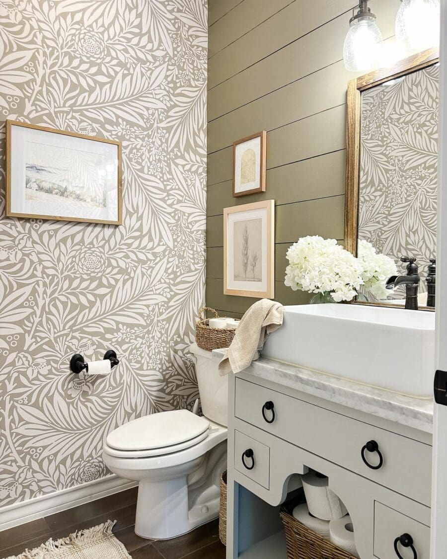 An elegant bathroom featuring beige floral wallpaper, a white porcelain toilet and a vanity with a marble top and black hardware. A olive green shiplap wall with framed art adds contrast, while a mirror with a golden frame reflects a bouquet of white hydrangeas. The scene is completed with a woven basket, a draped beige towel, and a neutral-toned rug on the floor.