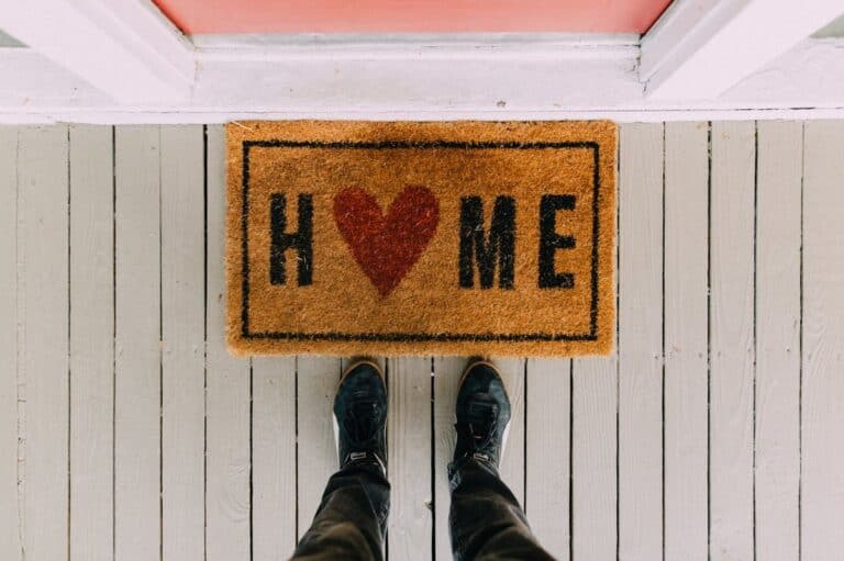 A person standing at a doorway with a welcoming doormat that spells 'HOME' with a heart symbol for the 'O', symbolising warmth and love in creating a healthy home environment.
