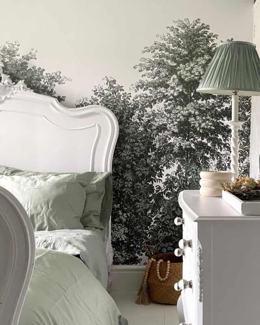 Elegant bedroom corner featuring a classic white headboard, soft green bedding, and a white nightstand with a lamp, set against a striking wallpaper mural of dense trees in a grayscale palette.