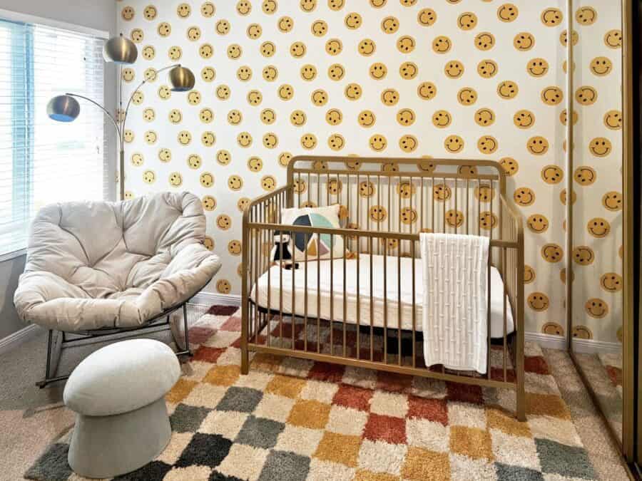 A playful nursery with smiley face-patterned wall, a gold crib, a cream Papasan chair with a footstool, a multicolored area rug, and a floor lamp with multiple round shades.
