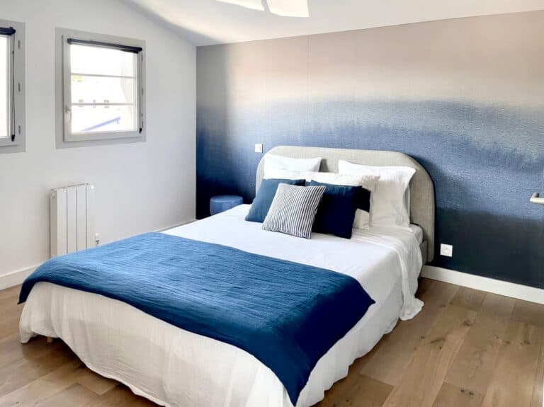 A bedroom designed to personalise your home, featuring a unique blue gradient accent wallpaper mural, white bed linen with a navy blue throw, complementing cushions and natural wooden flooring, illuminated by daylight from the windows.
