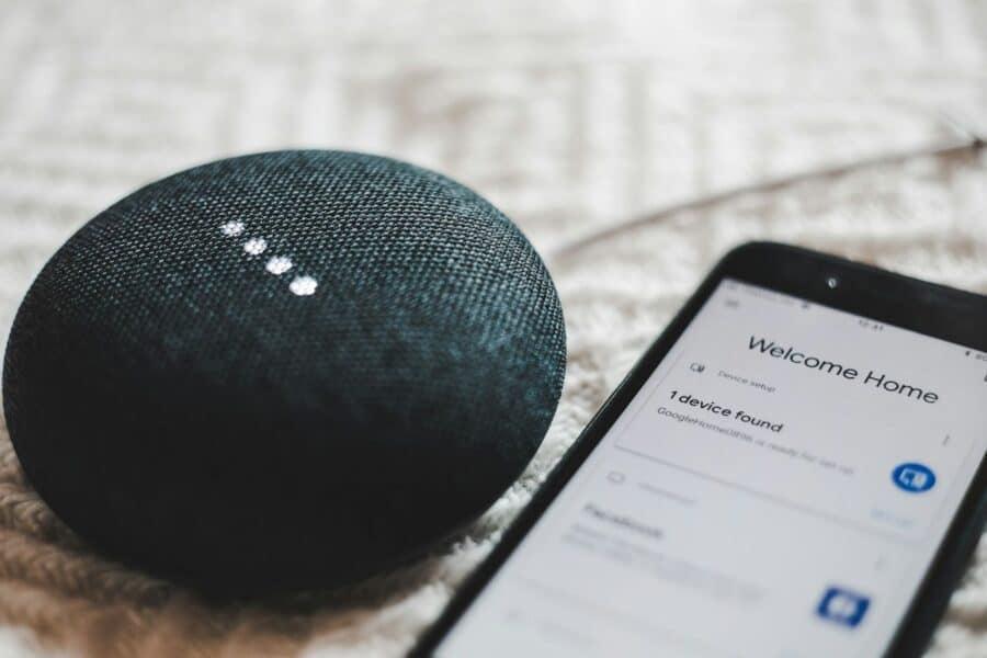 A smart speaker and a smartphone on a textured fabric surface, with the phone displaying a 'Welcome Home' message and recognising one device in a smart home setup, suggesting a connected and automated living environment.