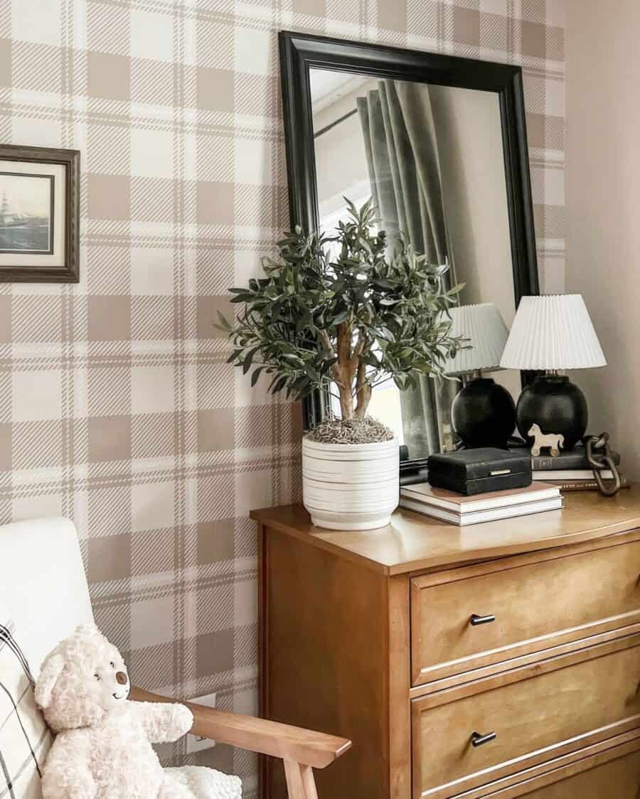 A cosy corner with beige plaid wallpaper, a wooden dresser topped with an olive tree in a white pot, books, a black lamp and a large mirror reflecting a curtained window. A plush teddy bear sits on a wooden chair.