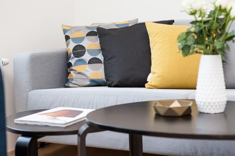 A cosy living room corner with a grey sofa adorned with patterned and yellow throw pillows, a white textured vase with fresh flowers on a round black coffee table, creating a comfortable and inviting home atmosphere.