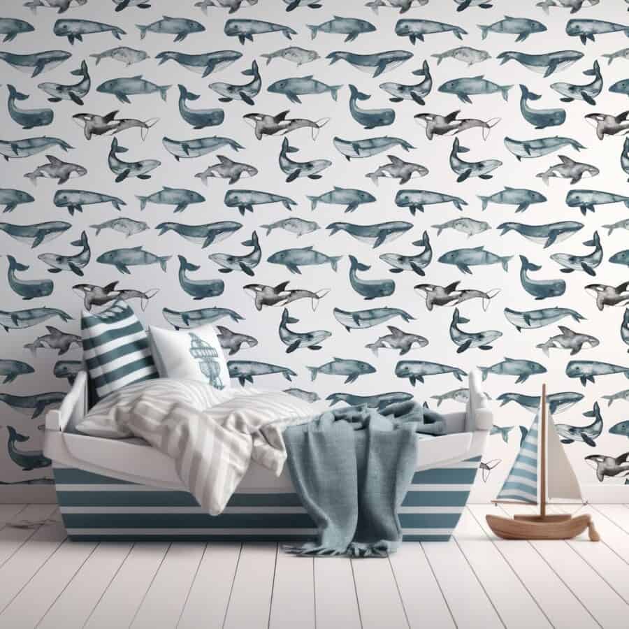 A cosy, nautical-themed bedroom corner featuring a bed with blue and white striped bedding, a lighthouse print pillow, a plush throw blanket and a model sailboat on a white wooden floor. The room is completed with whimsical wallpaper mural adorned with various blue whale illustrations.
