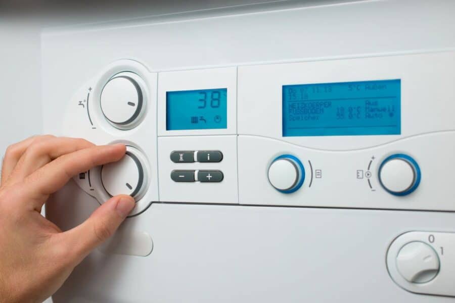 A person's hand is shown adjusting a dial on a modern boiler's control panel, with a digital display reading 38 degrees. The screen also shows various system statuses in German, emphasizing the importance of proper settings to increase the lifespan of your boiler.