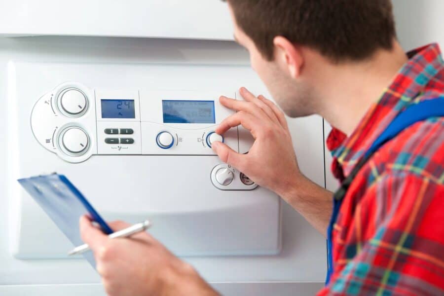 A technician in a red plaid shirt is focused on adjusting the settings on a boiler control panel, holding a clipboard for notes, indicating regular servicing to increase the lifespan of the boiler.