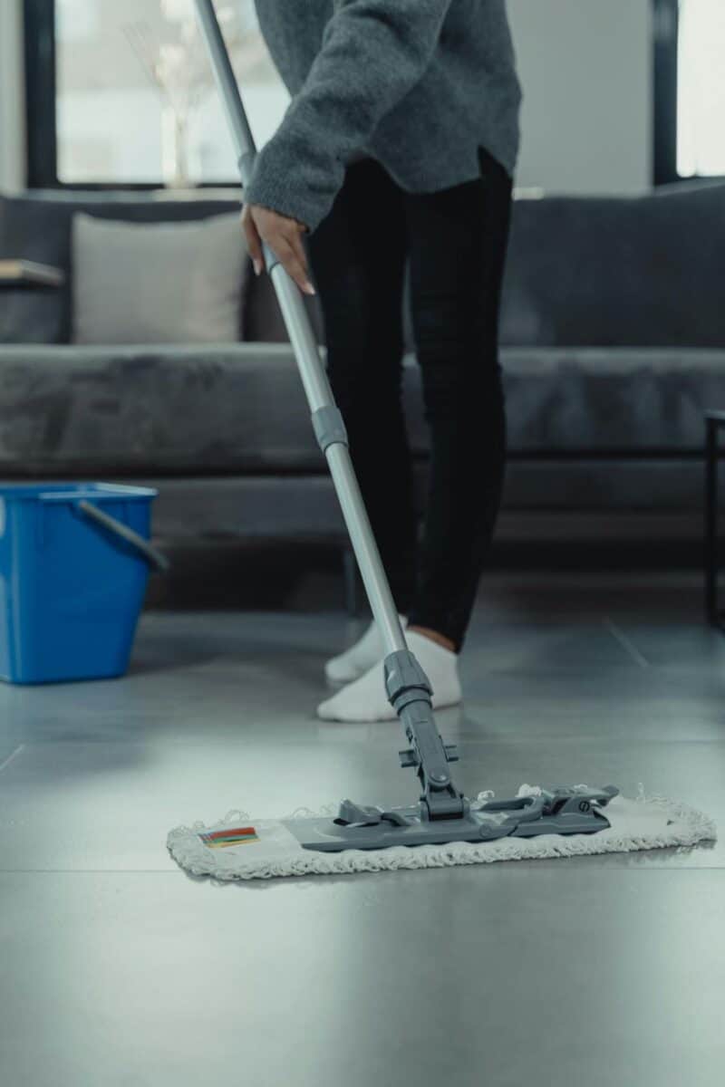 A person in a grey sweater mops a tiled floor, with a blue bucket placed nearby, indicating house cleaning in a living room setting. The mop and bucket suggest preparation for thorough cleaning.