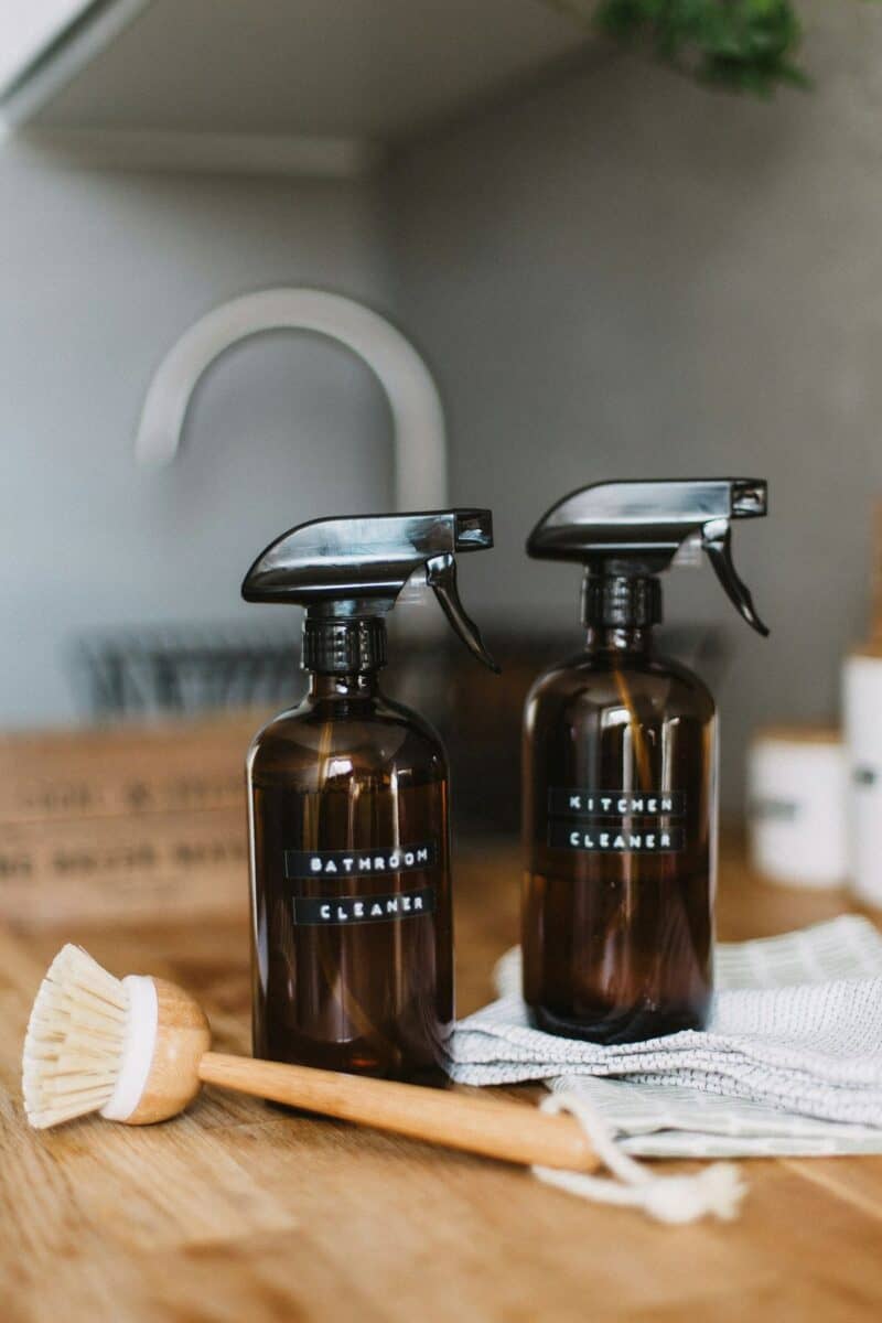 Two amber spray bottles labeled "Bathroom Cleaner" and "Kitchen Cleaner" sit on a wooden countertop next to a scrubbing brush and a cloth, suggesting homemade cleaning products for house cleaning.