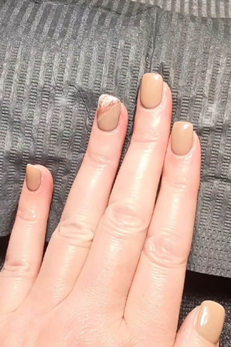 Here's a photo of my hand with a chic manicure featuring a taupe color on most nails and a unique white and gold marble effect on the ring finger, all set against a textured grey fabric backdrop.