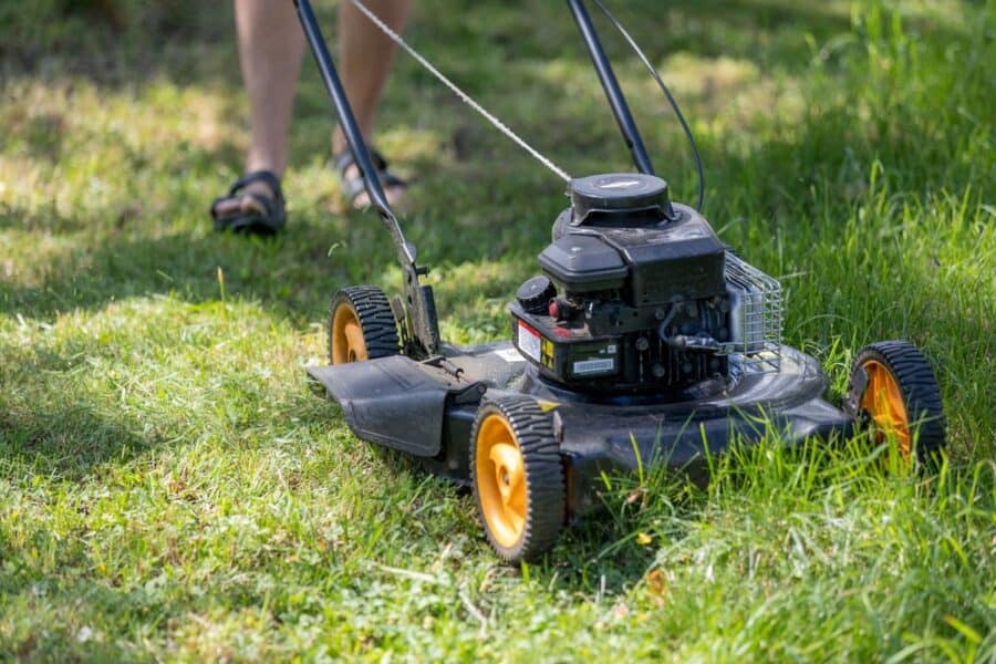 A person in sandals is using a gas-powered lawn mower to cut grass in a yard, highlighting outdoor maintenance and lawn care activities. The mower has black and orange features and is actively being pushed across the grassy area.