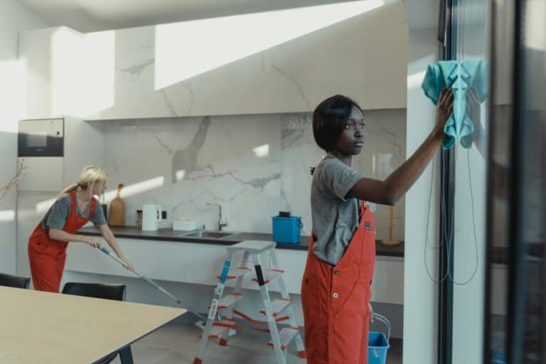 Two women wearing red overalls are diligently cleaning a house before moving out. One is wiping a large window with a blue cloth while the other mops the floor in a sleek, modern kitchen area, highlighting the importance of a thorough clean before vacating a property.