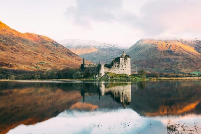 The ruins of Kilchurn Castle reflecting in the calm waters of Loch Awe, with the surrounding hills bathed in golden light. This image highlights the beauty and accessibility of travel in Scotland.